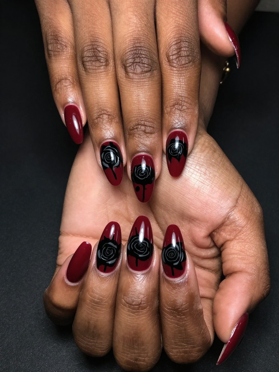 Top-down view of black feminine hands with coffin nails against a dark background. The nails are a glossy oxblood red. On each nail, a solid black silhouettes of dramatic roses cover the nails. The look is powerful, lush, and intensely romantic.