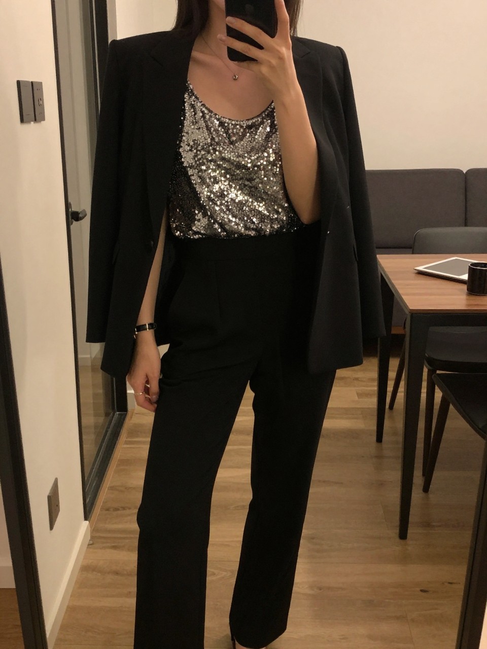 mirror selfie of a woman in a sequin tank and tuxedo pants, blazer open, cropped at NECK so face doesn't show, in a city apartment, warm moody lighting, iPhone photo quality.