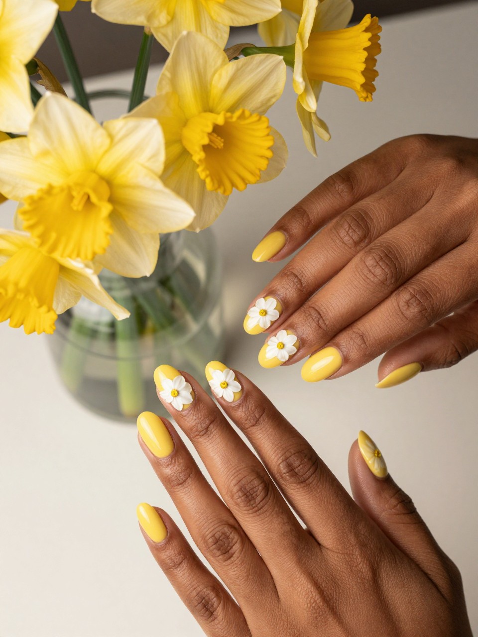 Overhead shot of black feminine hands with oval nails next to a bunch of daffodils vase. The nails are a glossy daffodil yellow. Each nail design features a single, detailed white narcissus flower with a small frilly cup center. The lighting is warm and spring-like.