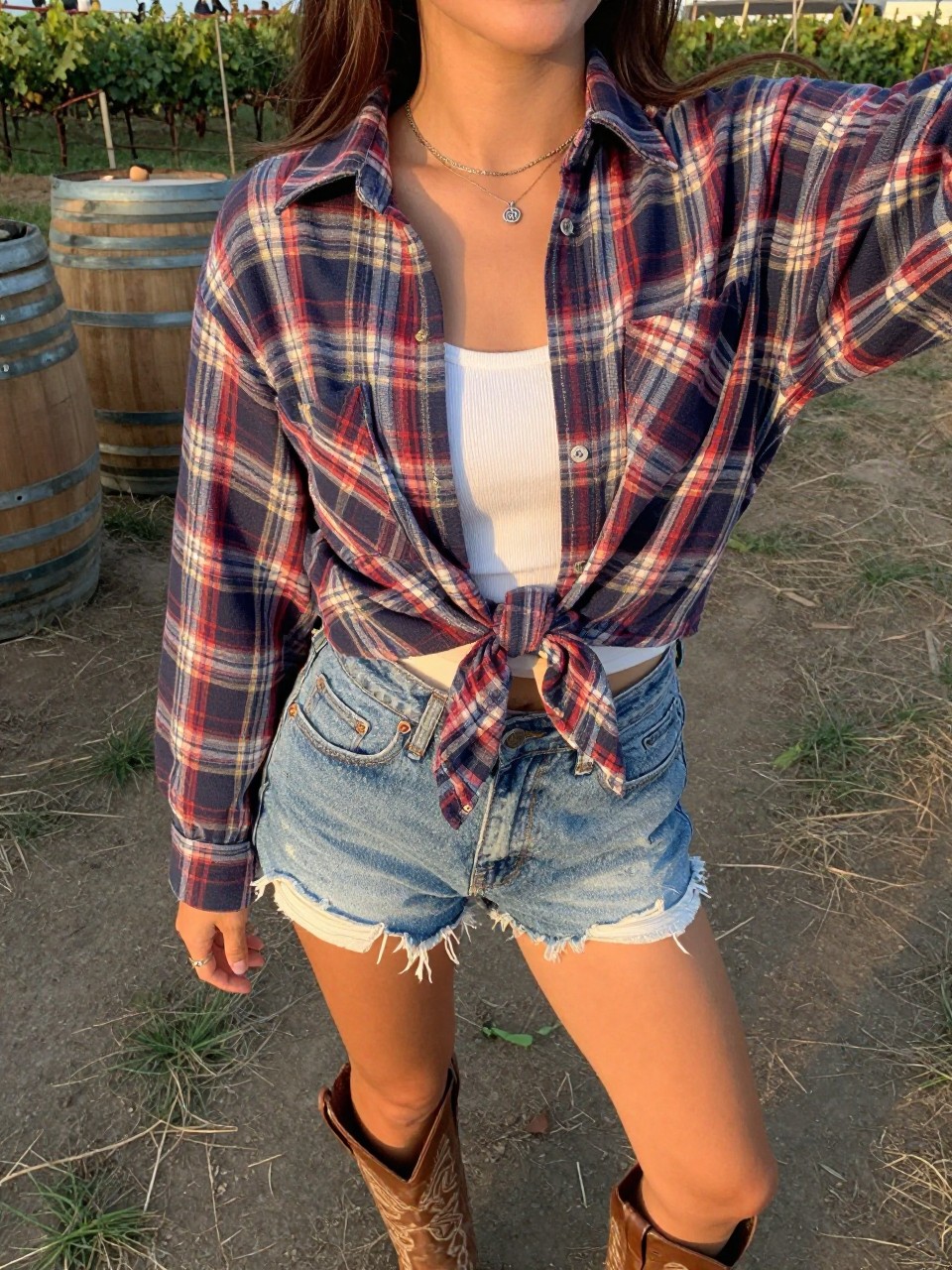 selfie of a woman in a plaid shirt tied over a white tank with denim shorts and cowboy boots, standing at an outdoor venue or ranch, cropped at NECK so face doesn't show, at a winery, evening golden hour, iPhone photo quality.