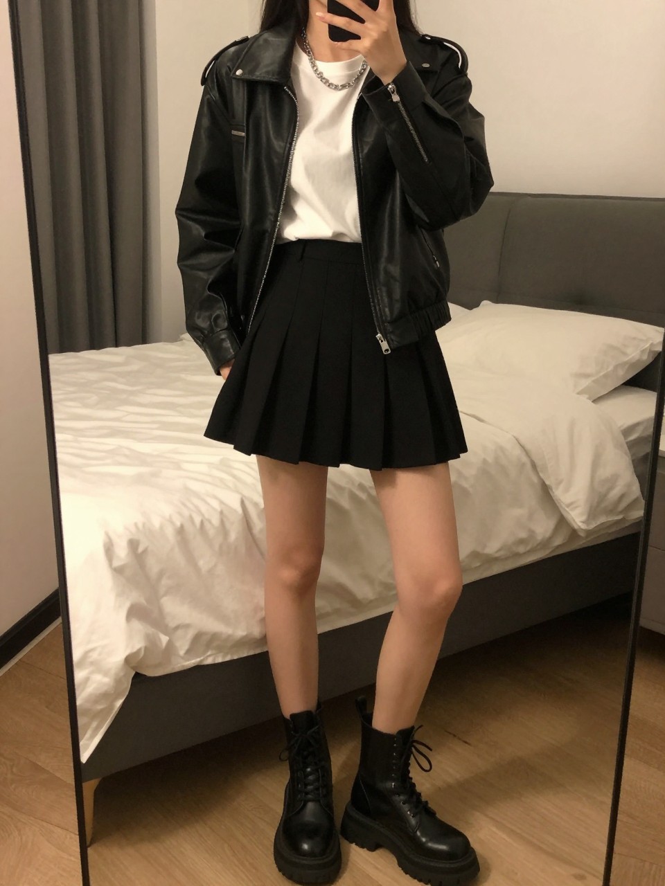 iPhone mirror selfie of a woman wearing a black leather jacket over a white tee tucked into a black pleated mini skirt with black chunky combat boots and a silver chain necklace, standing in an edgy pose with hands in pockets, cropped at NECK so face doesn't show, in a feminine-themed bedroom, warm moody lighting, iPhone photo quality.