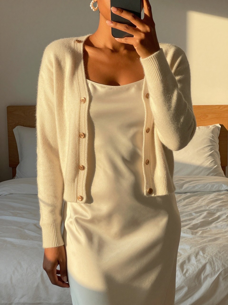 iPhone mirror selfie of a black woman wearing a cream cashmere cardigan over a simple tank with an ivory satin slip skirt and pearl earrings, one hand adjusting her cardigan, cropped at NECK so face doesn't show, in a feminine-themed bedroom, evening golden hour, iPhone photo quality.
