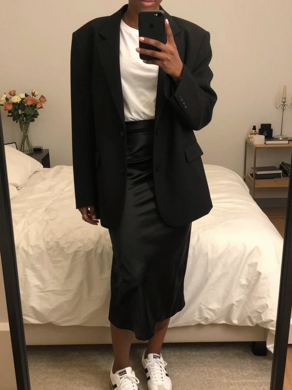 iPhone mirror selfie of a black woman wearing an oversized black blazer over a white tee tucked into a black satin midi skirt with white sneakers, one hand holding her phone at chest level, cropped at NECK so face doesn't show, in a feminine-themed bedroom, warm moody lighting, iPhone photo quality.