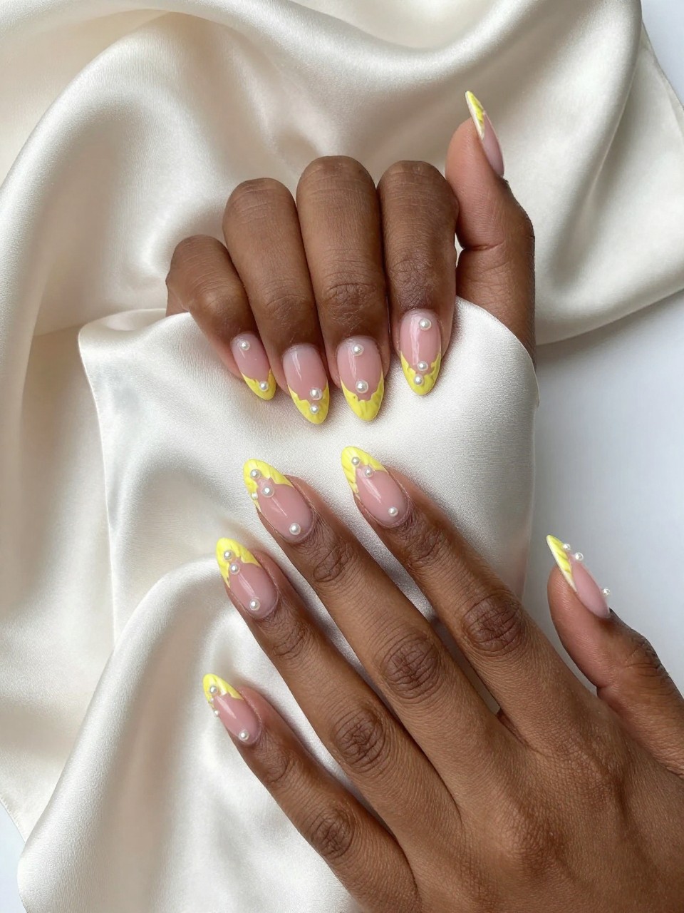 Overhead view of black feminine hands with almond nails draped over a silk scarf. The nails have a sheer pink base. Small yellow buttercup outlines dot each nail, each with a micro pearl in the center. Soft lighting highlights the delicate pearl textures.