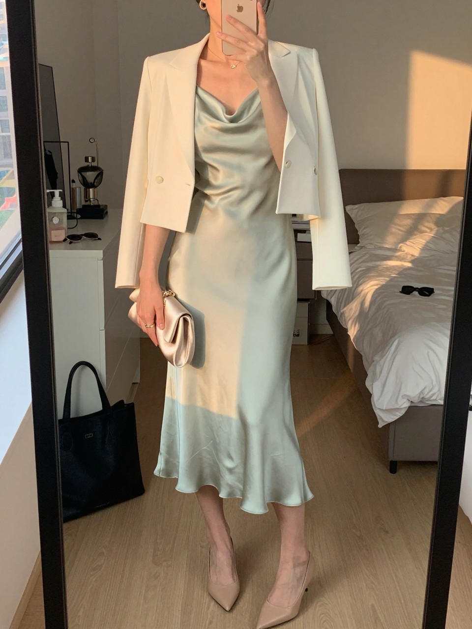 mirror selfie of a woman wearing a pale sage green satin midi dress with an ivory cropped tailored blazer, nude leather pointed-toe pumps, a champagne satin small clutch, and small simple gold hoop earrings, holding a phone in front of a mirror, cropped at NECK so face doesn't show, with a natural casual selfie pose, in a city apartment, evening golden hour, including small lived-in details like a tote bag on the floor, sunglasses on a dresser, or soft bedding in the background, latest iPhone photo quality.