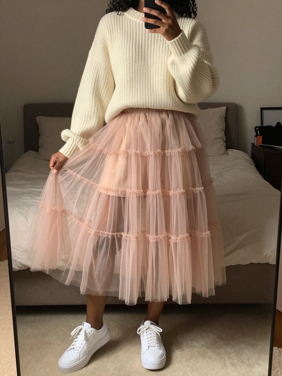 mirror selfie of a black woman wearing an oversized cream chunky sweater with a layered blush tulle fairy skirt and white platform sneakers, one hand holding her phone low by her thigh, cropped at NECK so face doesn't show, in a feminine-themed bedroom, warm moody lighting, iPhone photo quality.