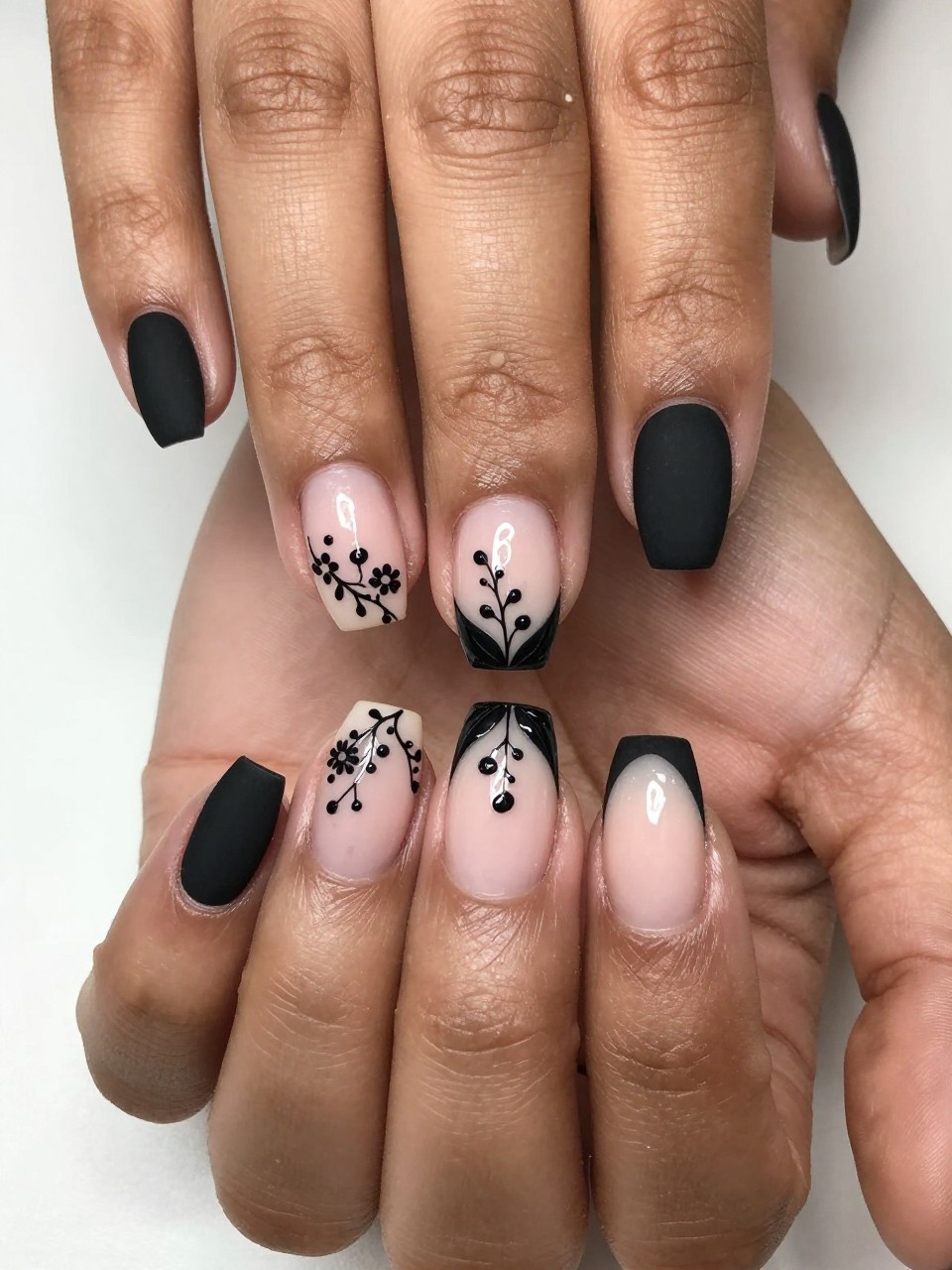 Close-up, top-down view of black feminine hands with natural, medium-length nails. The nails have a matte black French tip. Delicate black vines with micro leaves and tiny dot flowers trail from the tip down the nail bed. The look is intricate and sophisticated.