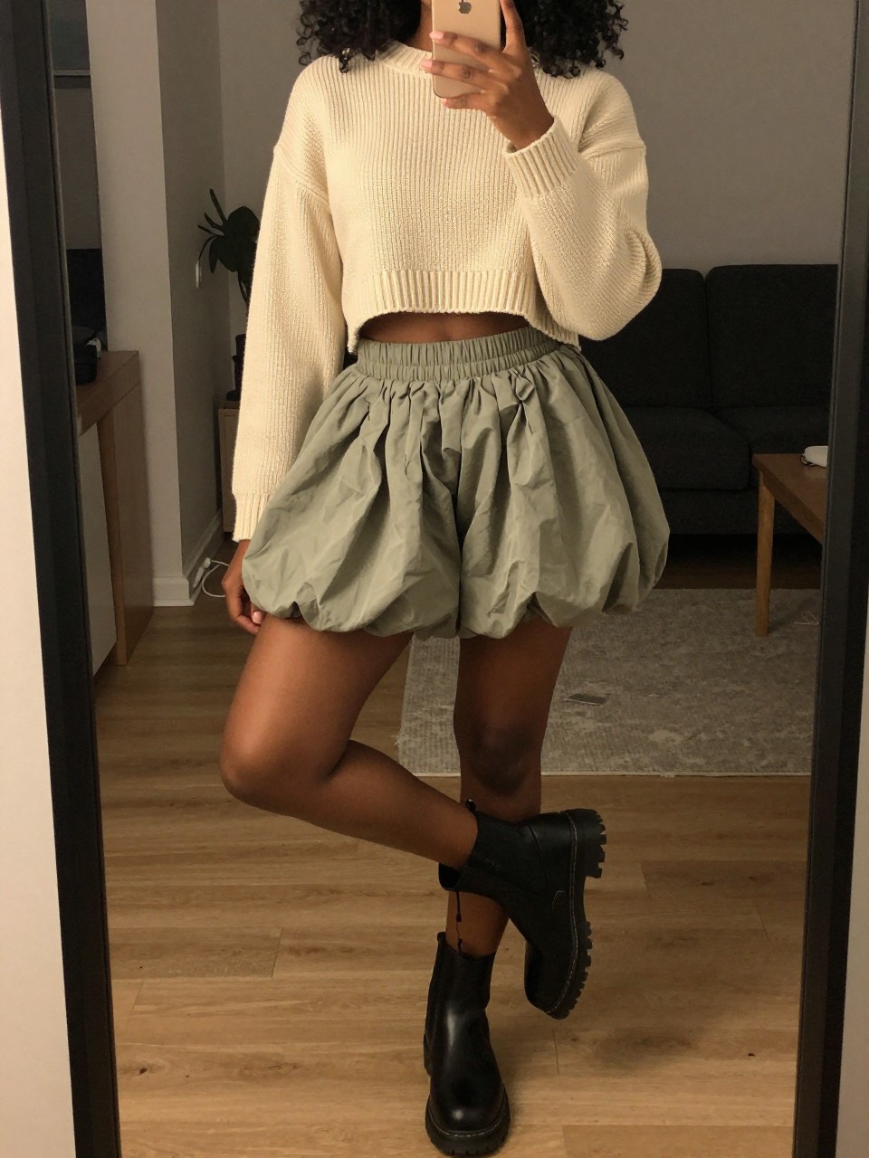 iPhone mirror selfie of a black woman in a cream cropped sweater and a sage green bubble skirt with lug-sole boots, standing with her weight on one leg, cropped at NECK so face doesn't show, in a city apartment, warm moody lighting, iPhone photo quality.