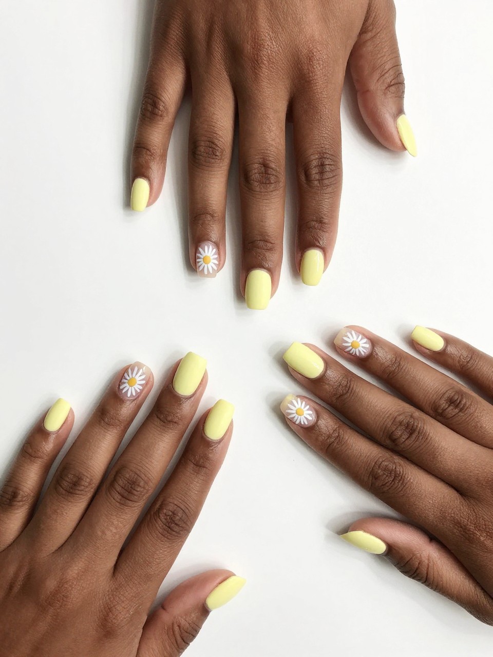 Top-down view of black feminine hands with neat, short nails on a white desk. The nails are mostly matte pastel yellow, but each has a slim crescent of bare nail along one side. A white daisy outline is painted inside that space. The look is architectural and cute.