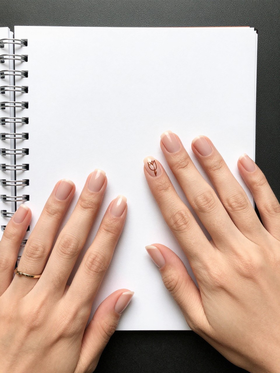 Overhead view of hands with pristine, natural nails on a white paper sketchpad. On one ring design, there is a minimalist brown outline of a tulip is drawn on the side of the nail. The rest are bare. The look is clean, intentional, and gallery-inspired.