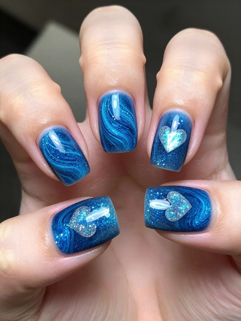Overhead view of hands with oval nails. The blue cat eye polish is swirled into a vortex pattern. At the vortex's center on each nail, the glitter forms a natural, shimmering heart. The look is like a miniature, romantic nebula.