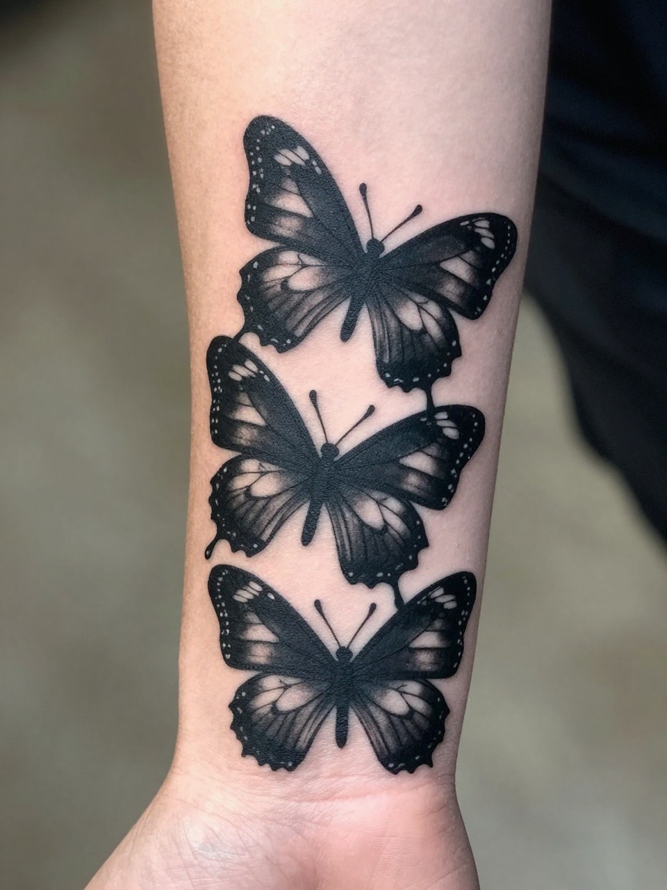Close-up of a wrist-to-forearm section tattoo: overlapping solid black butterfly silhouettes creating a rhythmic, graphic pattern. Stark contrast against the skin.