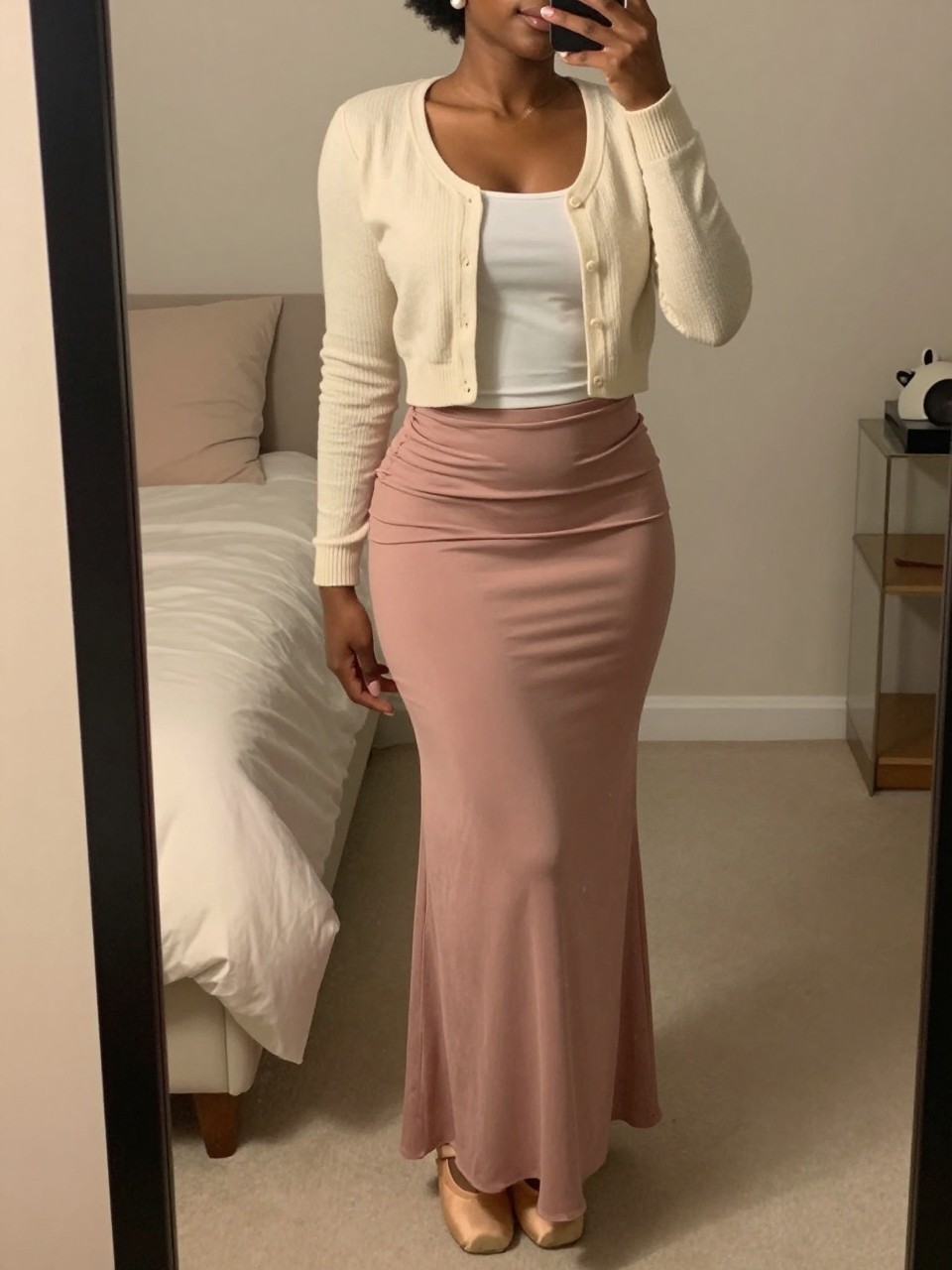 mirror selfie of a black woman wearing a cream cropped cardigan over a white tank top tucked into a dusty rose maxi skirt with nude ballet flats and pearl stud earrings, standing in a soft feminine pose, cropped at NECK so face doesn't show, in a feminine-themed bedroom, warm moody lighting, iPhone photo quality.