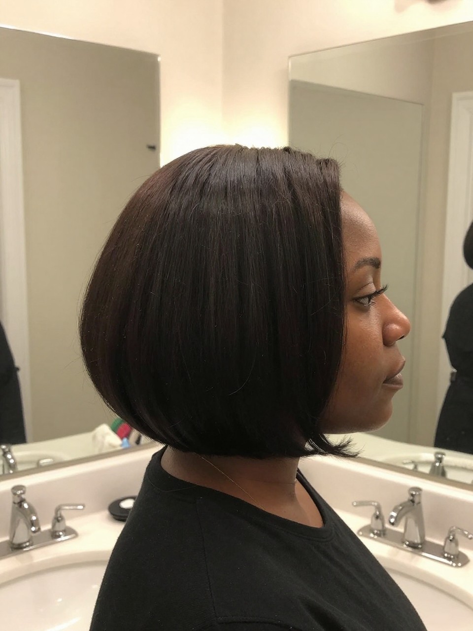 Photo of a black woman wearing a natural bob with shrinkage, side profile view showing the rounded compact shape, setting is a bathroom vanity mirror with soft indoor lighting, containing a diffuser attachment visible, iPhone photo quality.