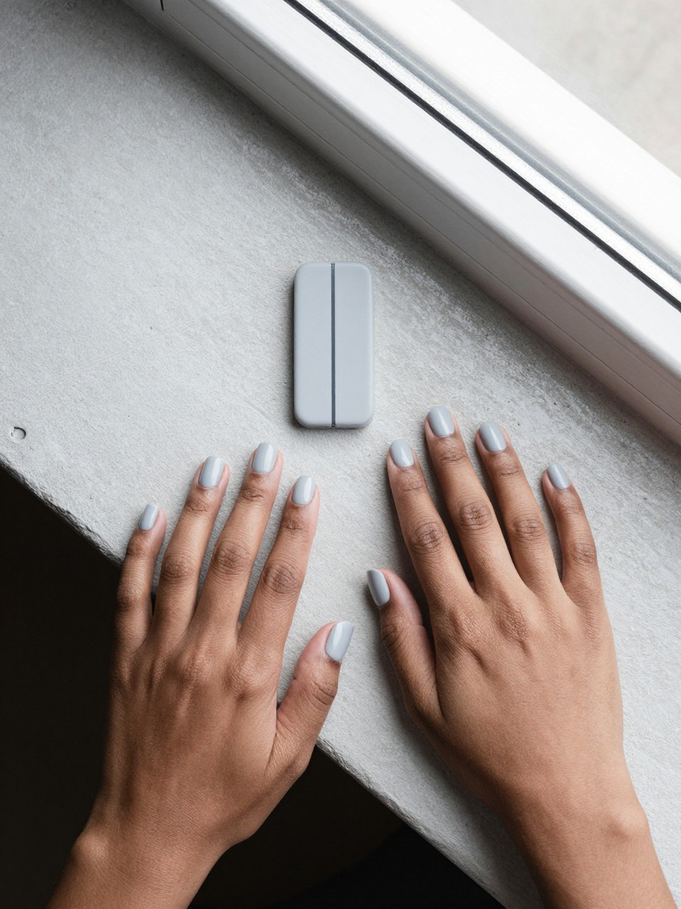 Overhead view of black feminine hands with short, straight-edged nails posed on a concrete windowsill. The base color is a light matte grey. A thin, dark matte grey line runs vertically down one side of each nail. The composition is graphic and clean, with bright natural light.