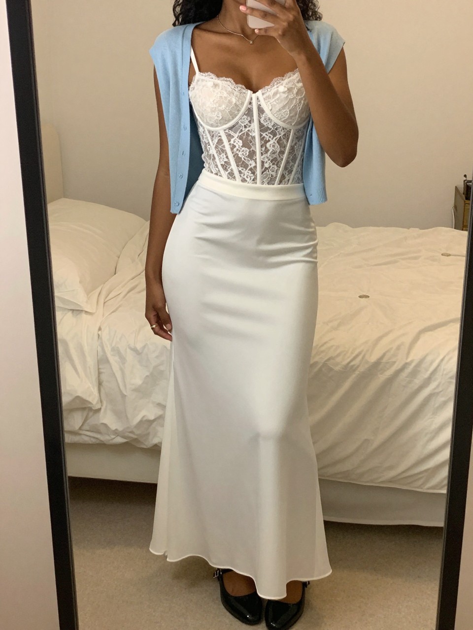 mirror selfie of a black woman wearing a white lace corset top with a high-waisted white maxi skirt, a baby blue cardigan draped over her shoulders, wearing patent Mary Janes, standing in a soft pose, cropped at NECK so face doesn't show, in a feminine-themed bedroom, warm moody lighting, iPhone photo quality.