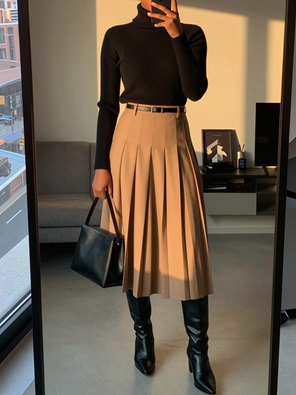 iPhone mirror selfie of a black woman wearing a black fine-knit turtleneck tucked into a camel pleated midi skirt with black leather knee-high boots and a thin black belt, holding a structured black shoulder bag, standing in an elegant pose, cropped at NECK so face doesn't show, in a city apartment, evening golden hour, iPhone photo quality.