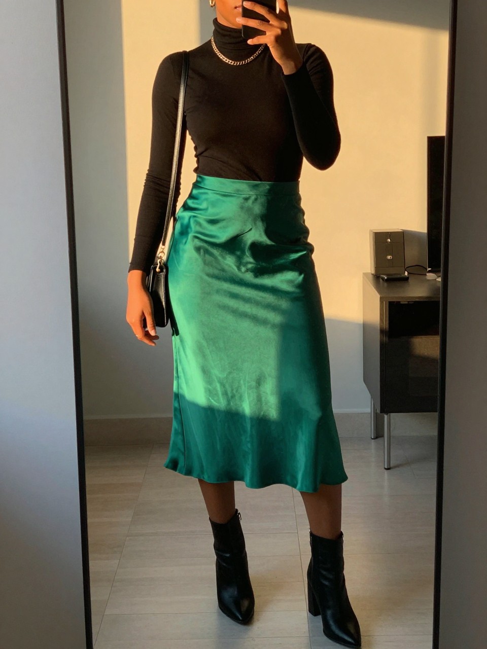 iPhone mirror selfie of a black woman wearing a black turtleneck tucked into an emerald green satin midi skirt with black block-heel ankle boots and a gold chain necklace, holding a black crossbody bag, standing in an elegant pose, cropped at NECK so face doesn't show, in a city apartment, evening golden hour, iPhone photo quality.