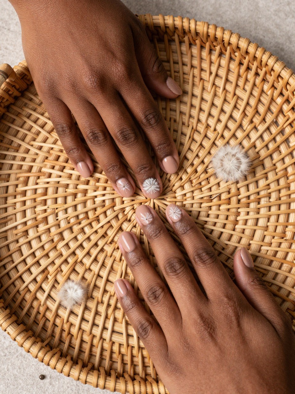 Overhead shot of black feminine hands with short nails on a woven rattan tray. The nails are a matte light biscuit brown. A soft white dandelion puff is painted on each nail, with a few drifting seed lines. The ambiance is rustic and gently artistic.