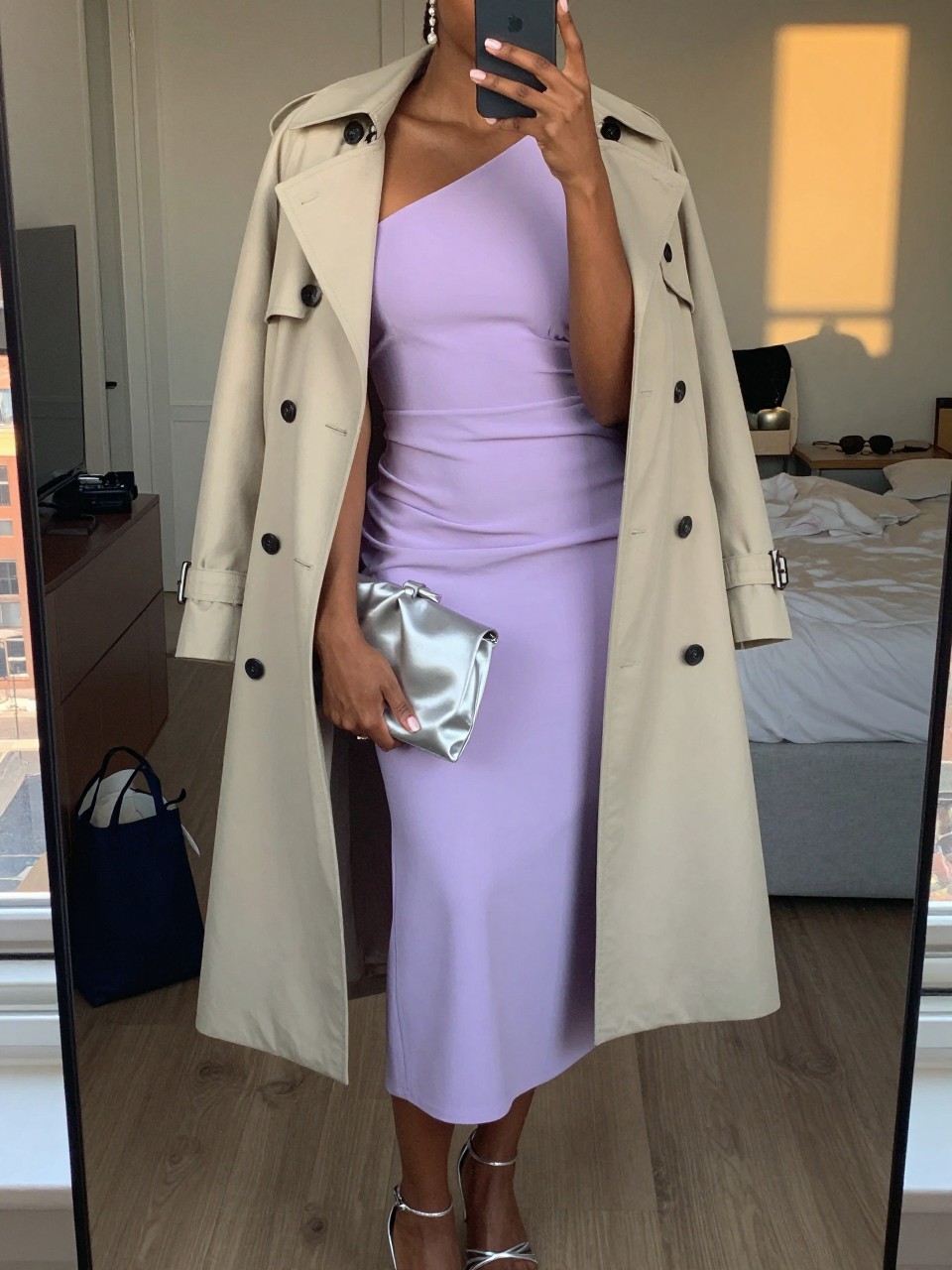 mirror selfie of a black woman wearing a soft lavender crepe one-shoulder midi dress with a stone beige light trench coat, silver strappy heels, simple crystal drop earrings, and a silver satin small clutch, holding a phone in front of a mirror, cropped at NECK so face doesn't show, with a natural casual selfie pose, in a city apartment, evening golden hour, including small lived-in details like a tote bag on the floor, sunglasses on a dresser, or soft bedding in the background, latest iPhone photo quality.