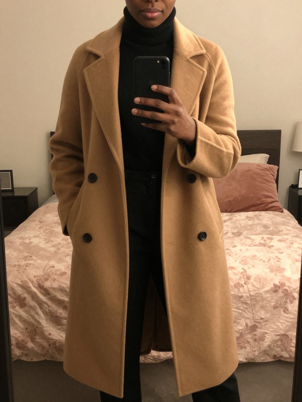 mirror selfie of a Black woman wearing a camel coat over a black turtleneck and trousers, standing with her weight shifted and phone low, cropped at NECK so face doesn't show, in a feminine-themed bedroom, warm moody lighting, iPhone photo quality.