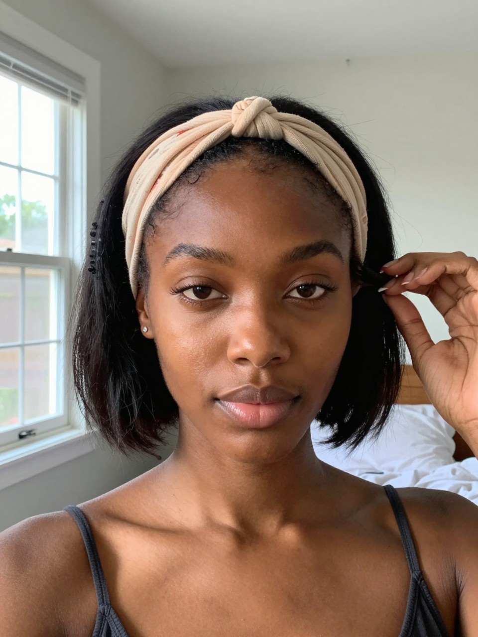 Photo of a black woman wearing a textured bob with a knotted headband, front-facing view showing the headband at her hairline, setting is a sun-drenched bedroom with window light, containing her hand adjusting a curl, iPhone photo quality.