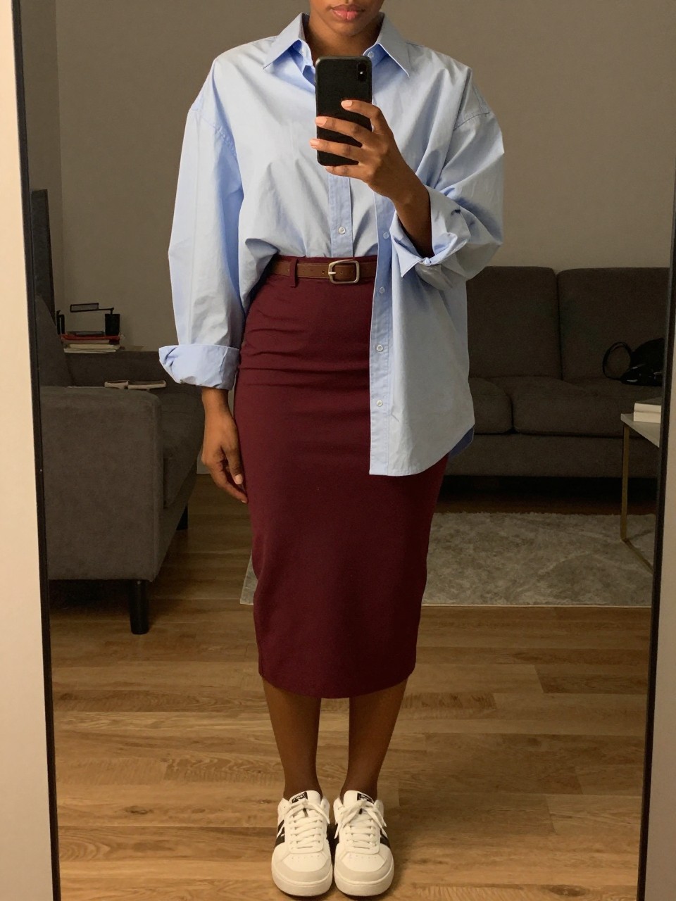 mirror selfie of a black woman wearing an oversized light blue button-down over a maroon pencil skirt cinched with a brown belt and white sneakers, holding her phone at chest level, cropped at NECK so face doesn't show, in a city apartment, warm moody lighting, iPhone photo quality.