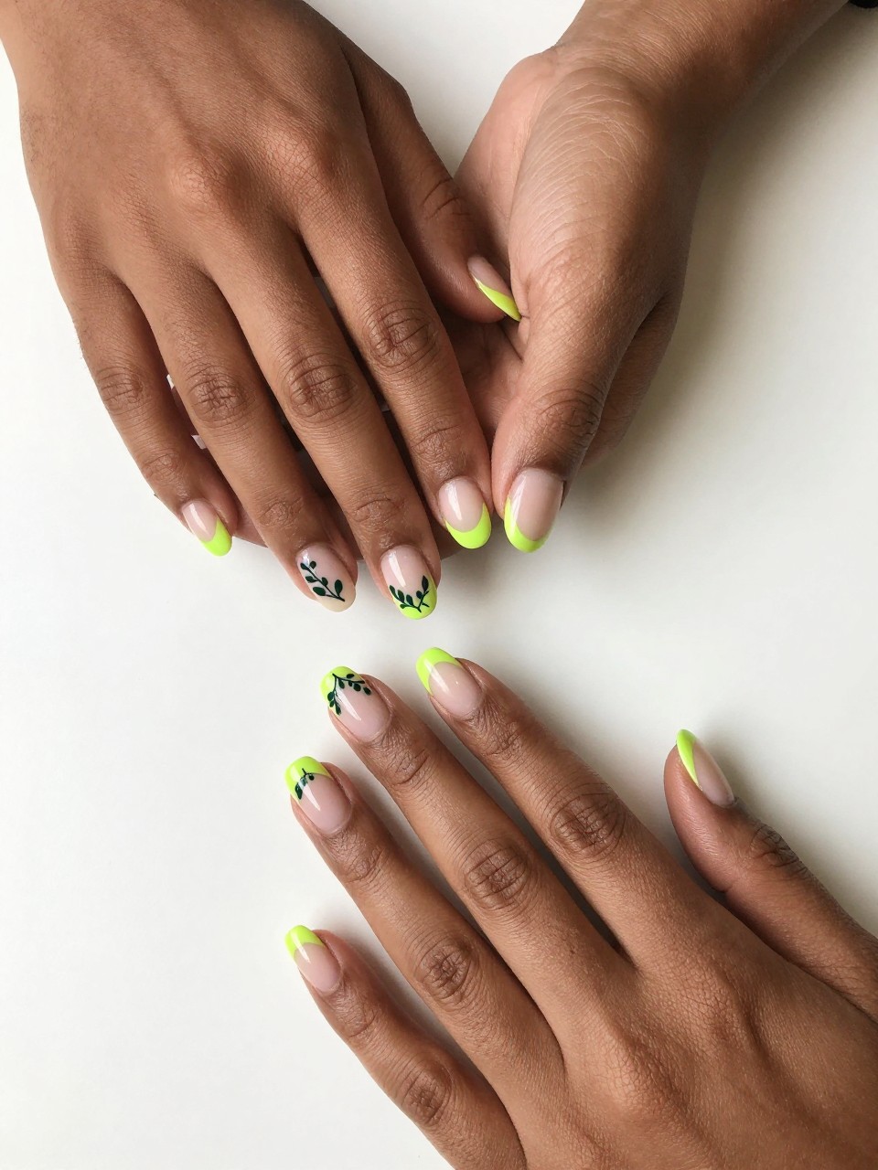 Top-down view of black feminine hands with natural, medium-length nails on a white desk. The nails have a bright chartreuse French tip. Delicate dark green vines with small leaves is designed on the tip down the nail bed. The look is crisp and botanical.