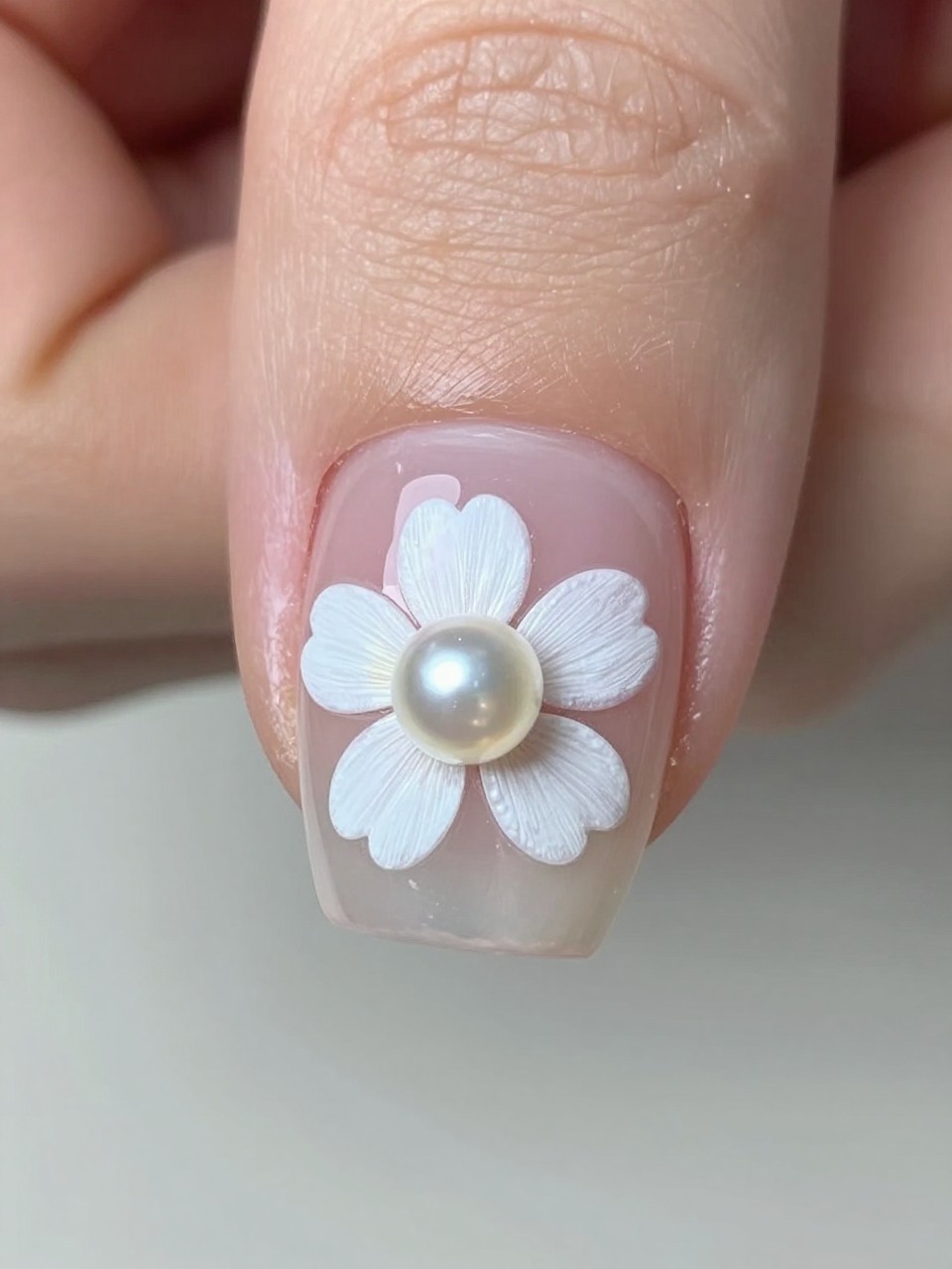 Close-up, bird's-eye view of a single nail. The nail is clear and glossy. A small white flower is painted, with one micro pearl placed perfectly in its center. The lighting shows the slight shadow of the pearl, highlighting the texture.