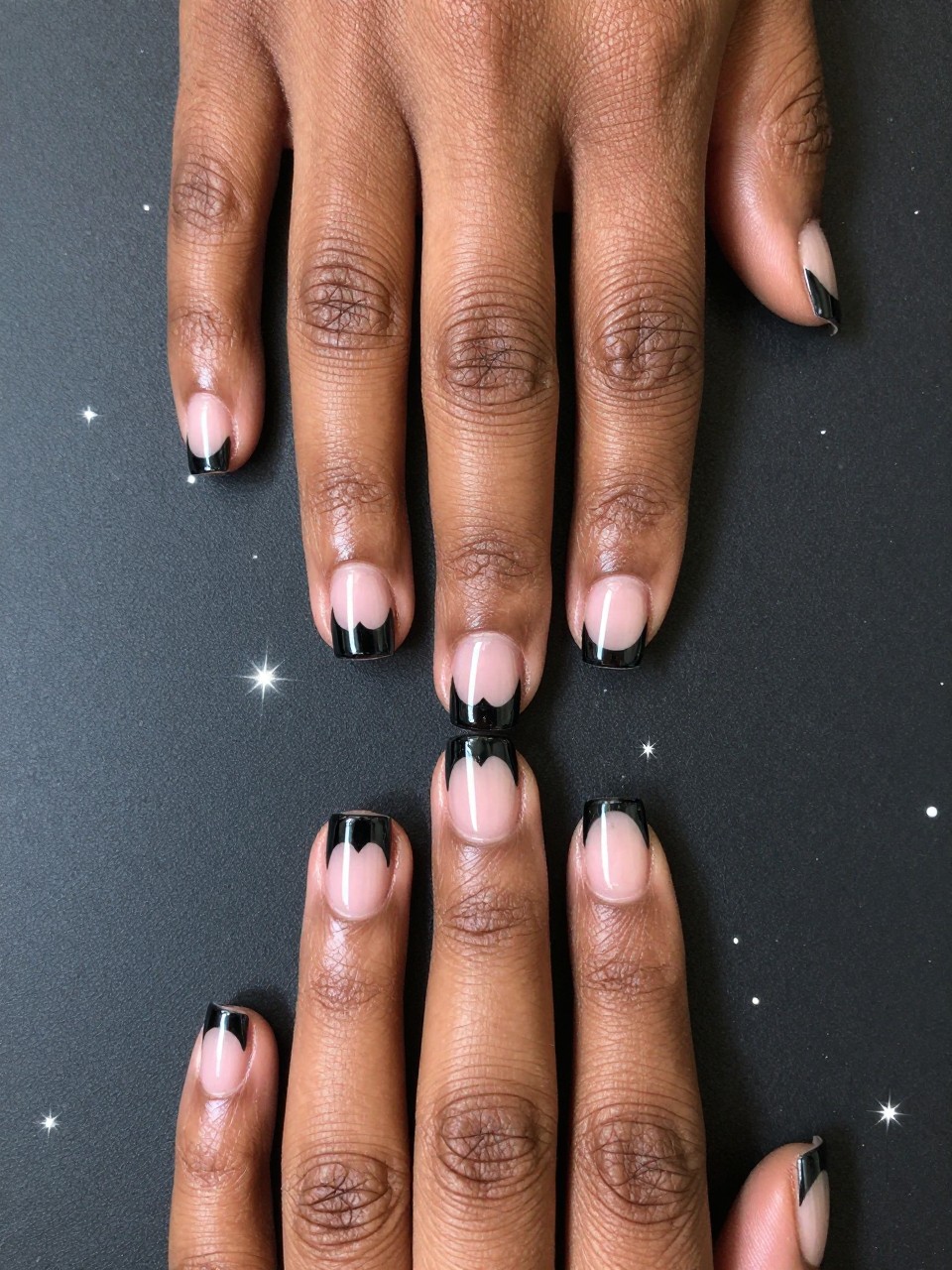 Top-down view of black feminine hands with short, squared nails against a starry backdrop. The nails are a black cat eye with a strong magnetic line. A clean, heart-shaped cut-out at the tip of each nail reveals the bare nail underneath. The look is cool and architectural.