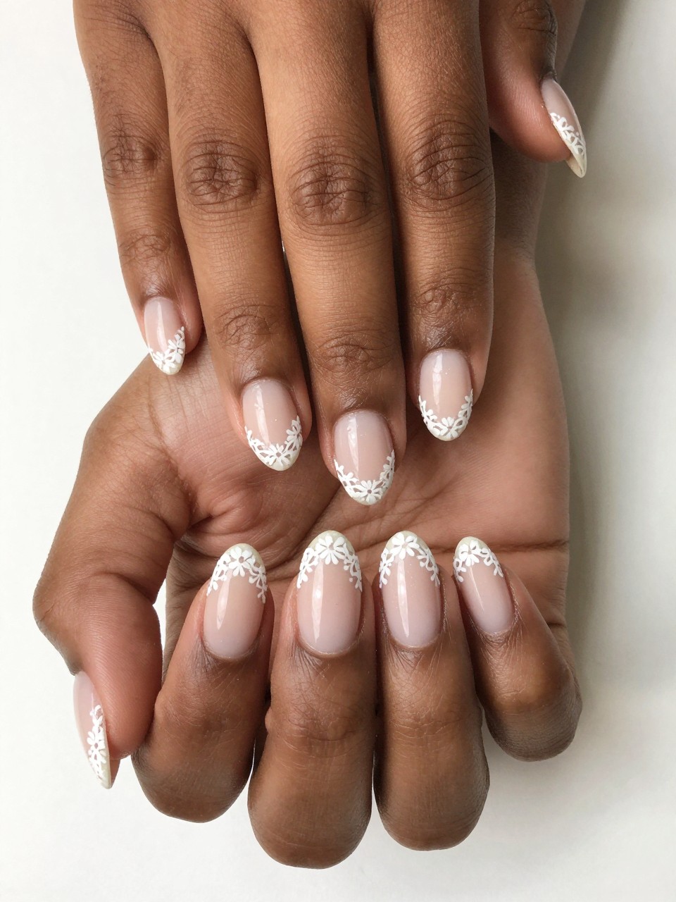 Bird's-eye view of black feminine hands with oval nails. The nails have a sheer nude base. The very edge of each tip is detailed with a precise white lace pattern featuring micro flowers. The look is vintage-inspired, dainty, and perfectly finished.