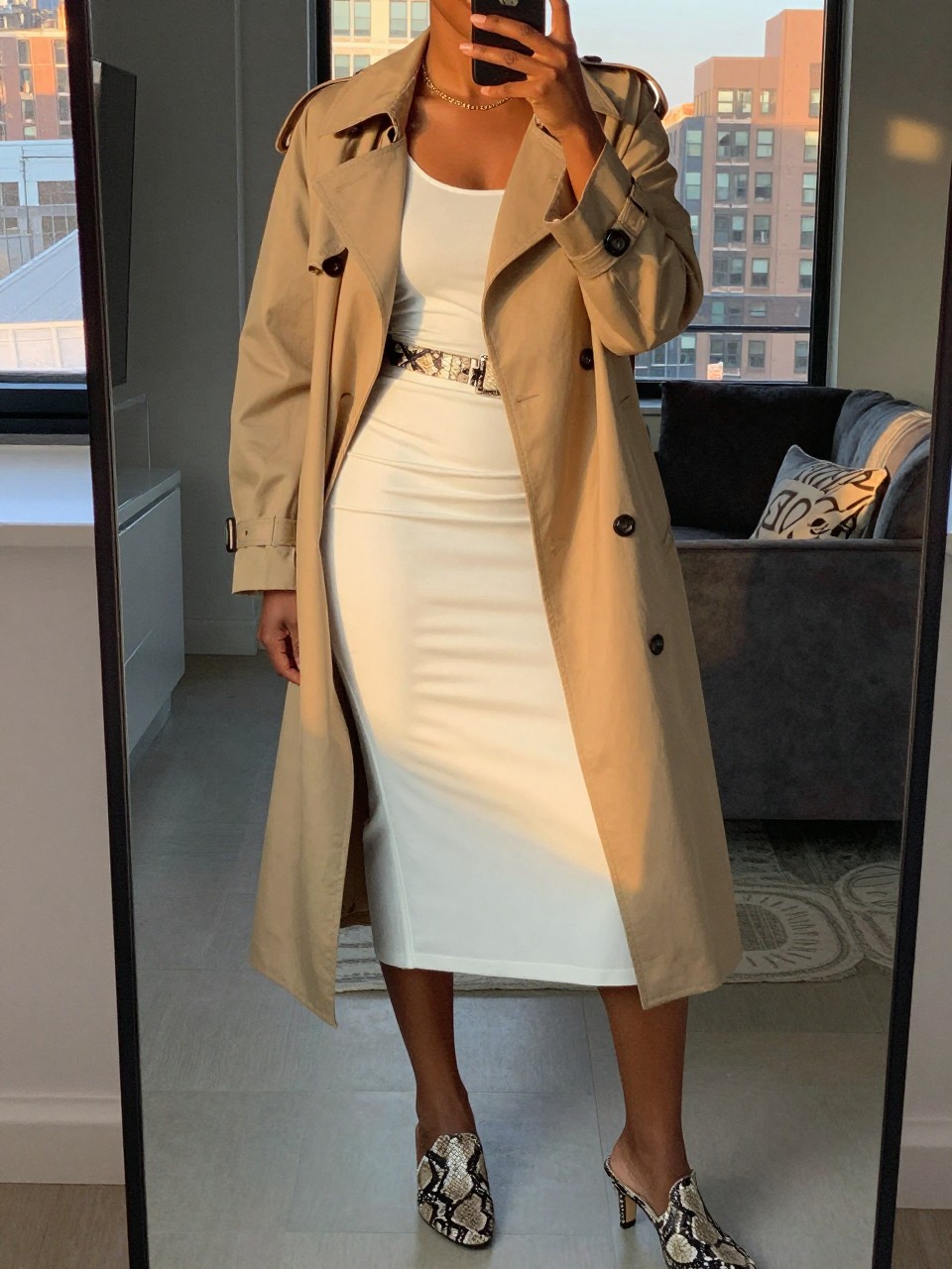 mirror selfie of a black woman wearing a tan trenchcoat over a white tank top and white midi skirt, cinched with a snake-print belt, with snake-print mules, standing in a city-ready pose, cropped at NECK so face doesn't show, in a city apartment, evening golden hour, iPhone photo quality.