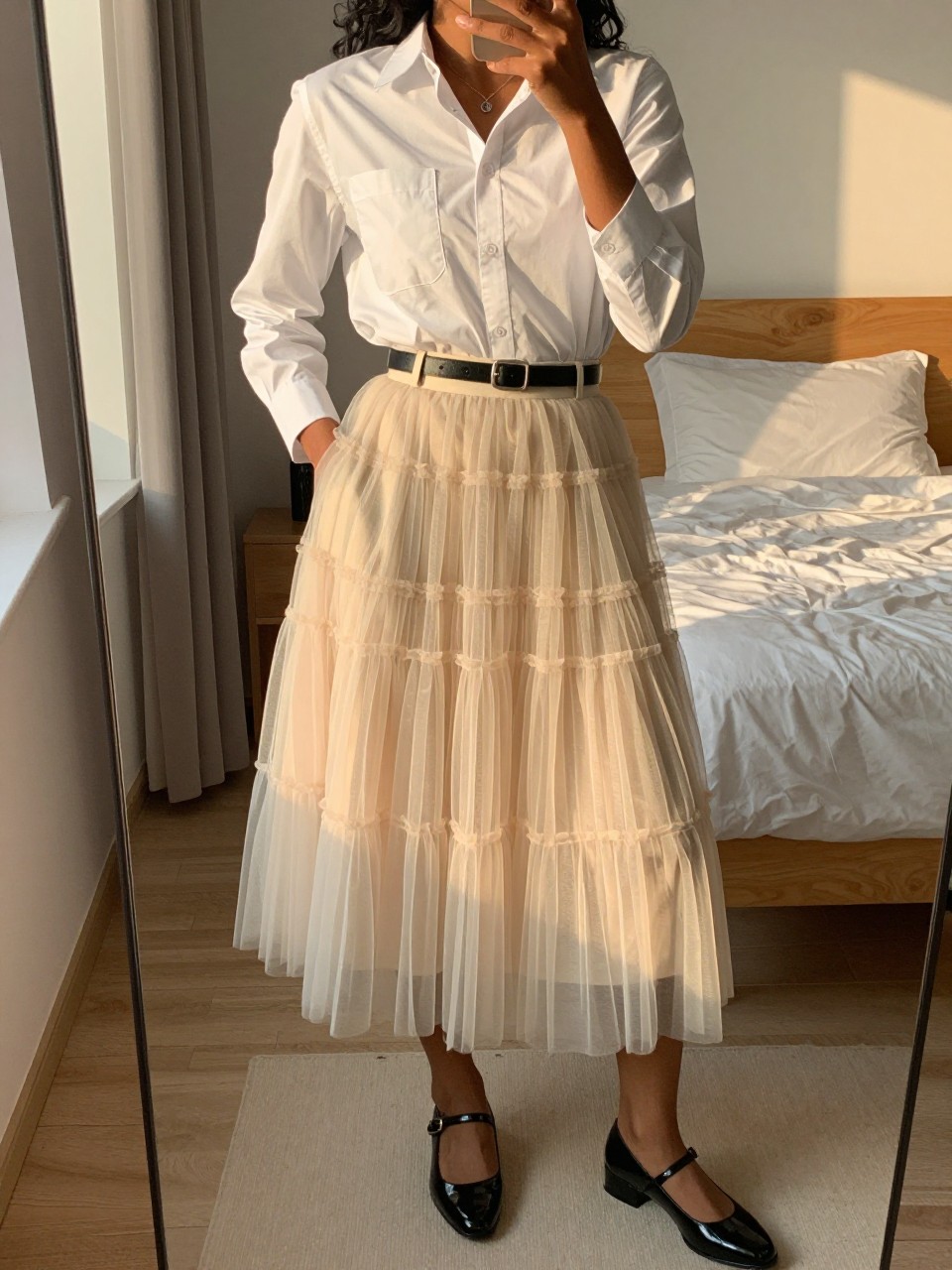 mirror selfie of a black woman wearing a white button-down shirt tucked into a cream layered tulle fairy skirt with black patent Mary Janes and a thin black belt, one hand in her pocket, cropped at NECK so face doesn't show, in a feminine-themed bedroom, evening golden hour, iPhone photo quality.