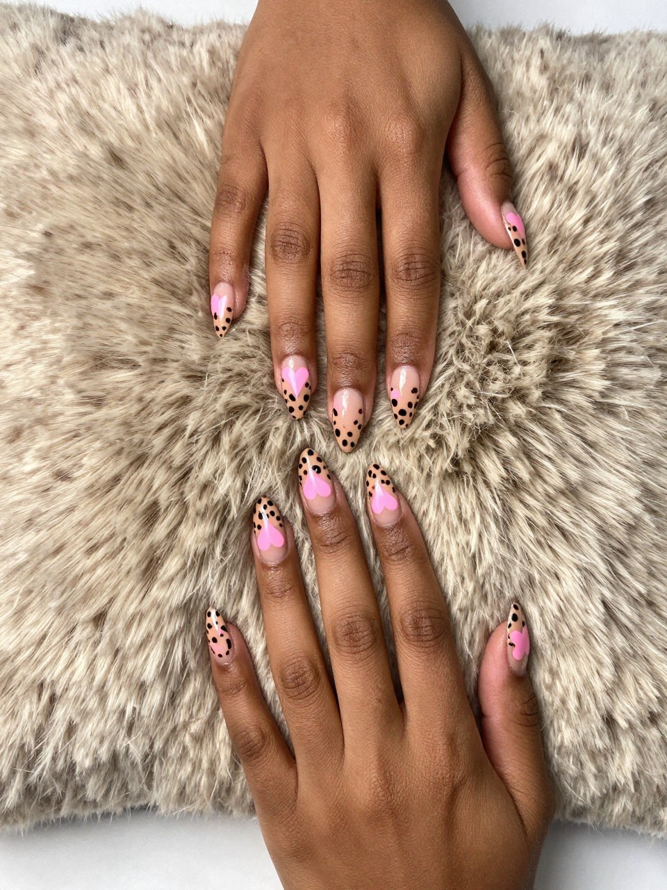 Bird's-eye view of black feminine hands with almond nails resting on a faux fur pillow. The nails have a tan base with small pink hearts forming a leopard-print pattern. The look is trendy, playful, and unexpectedly cute.