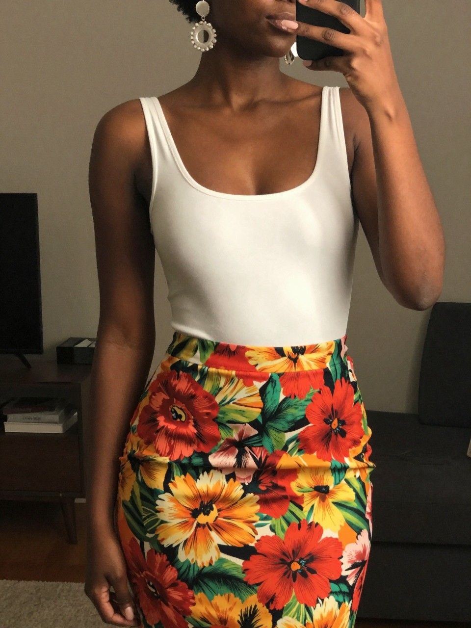 iPhone mirror selfie of a black woman in a white bodysuit and a vibrant floral skirt with statement earrings, one hand gently touching her earring, cropped at NECK so face doesn't show, in a city apartment, warm moody lighting, iPhone photo quality.