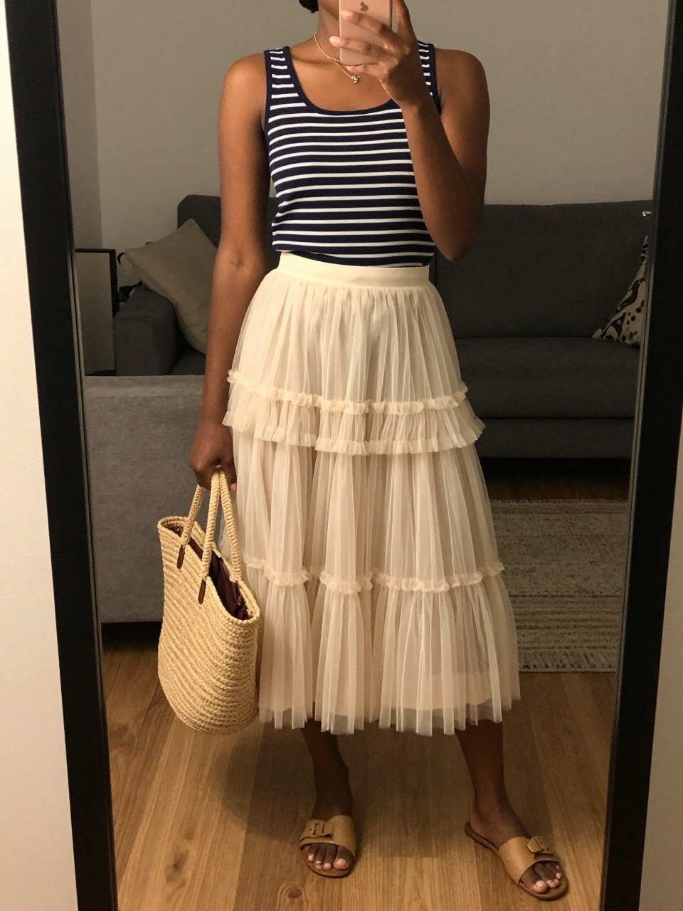 mirror selfie of a black woman wearing a navy striped boatneck top tucked into a cream layered tulle fairy skirt with flat tan sandals and a straw tote, one hand holding her phone at her side, cropped at NECK so face doesn't show, in a city apartment, warm moody lighting, iPhone photo quality.