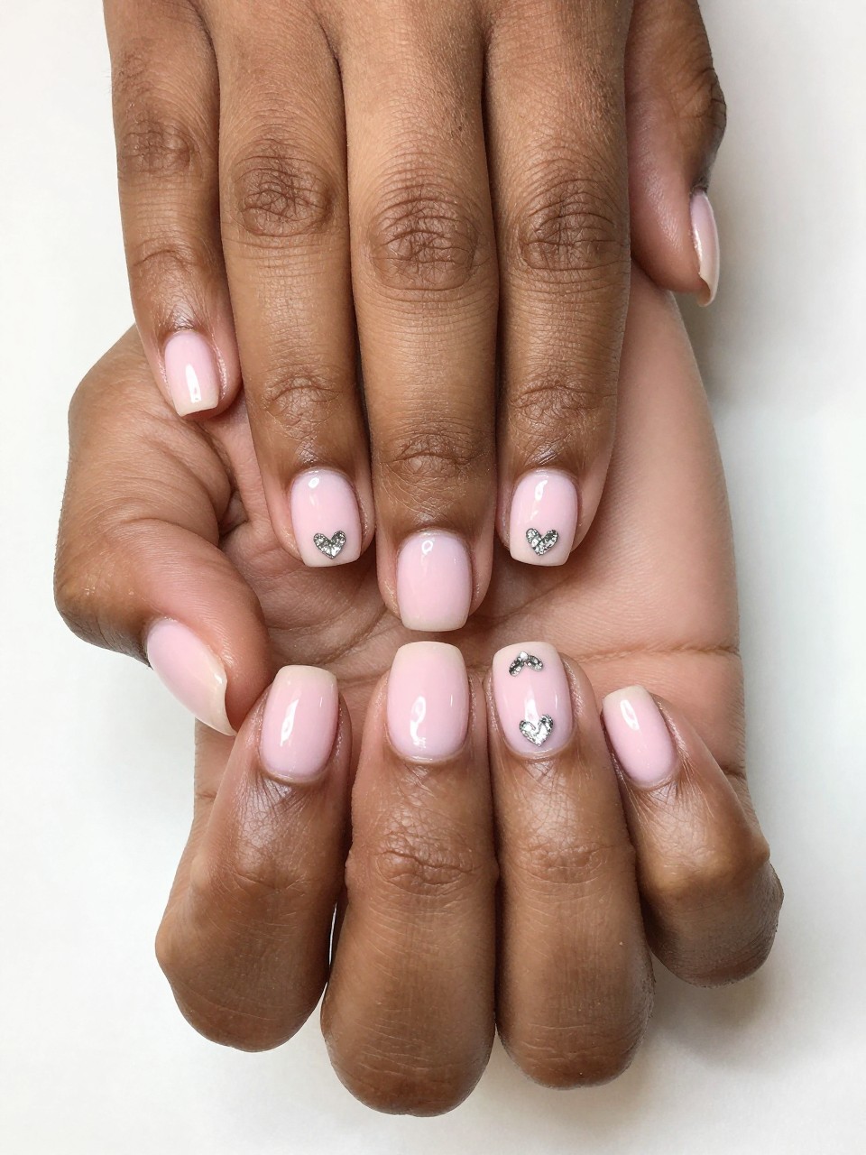 Bird's-eye view of black feminine hands with short, rounded nails. The nails are a creamy baby pink. At the very tip of each nail, a small, upside-down silver cat eye heart shimmers. The setting is bright and feminine.
