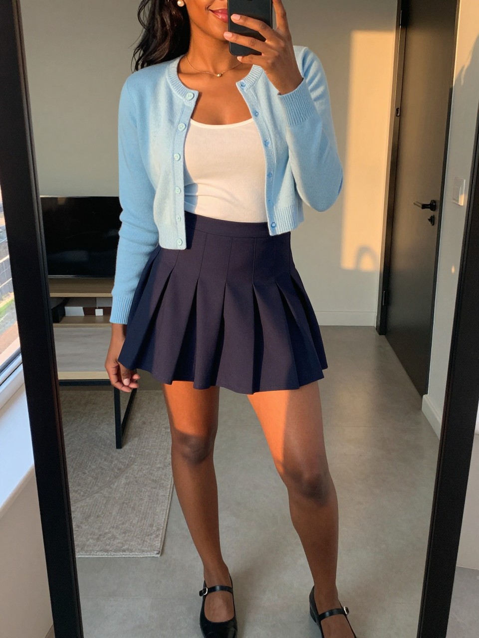 iPhone mirror selfie of a black woman wearing a baby blue cropped cardigan over a white tank top tucked into a navy pleated mini skirt with black Mary Jane flats and pearl stud earrings, standing in a sweet polished pose, cropped at NECK so face doesn't show, in a city apartment, evening golden hour, iPhone photo quality.