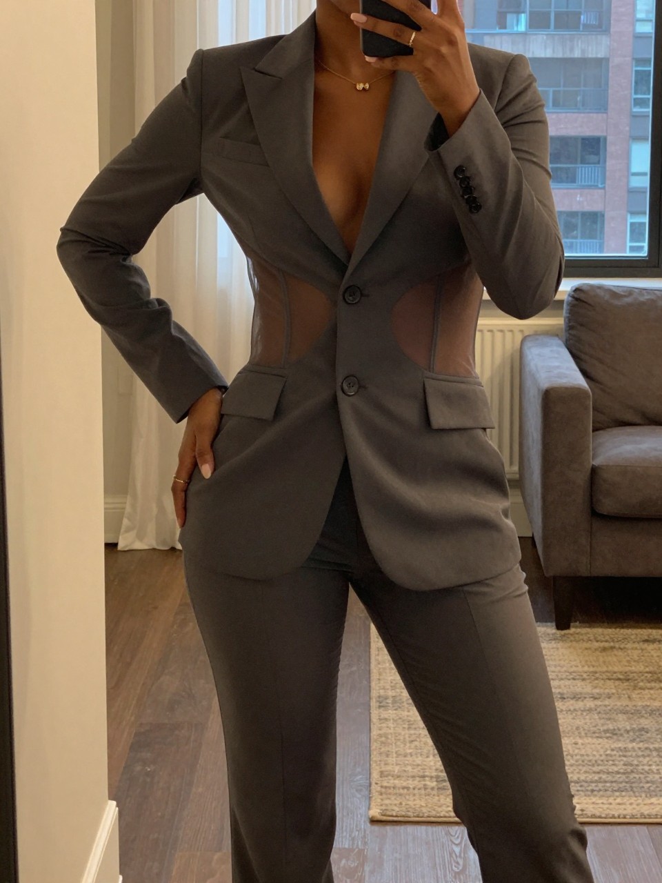mirror selfie of a black woman in a pantsuit with sheer panels, hand on hip, cropped at NECK so face doesn't show, in a city apartment, warm moody lighting, iPhone photo quality.