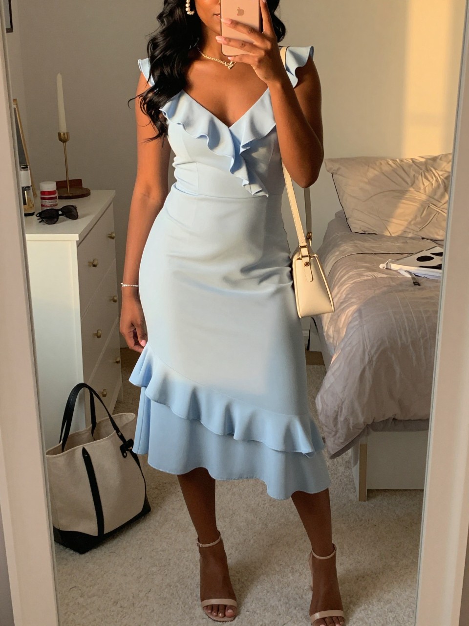 mirror selfie of a black woman wearing a soft sky blue ruffle midi dress with a cream light wrap, nude heeled sandals, a pearl hair clip in loose waves, and an ivory mini shoulder bag, holding a phone in front of a mirror, cropped at NECK so face doesn't show, with a natural casual selfie pose, in a feminine-themed bedroom, evening golden hour, including small lived-in details like a tote bag on the floor, sunglasses on a dresser, or soft bedding in the background, latest iPhone photo quality.