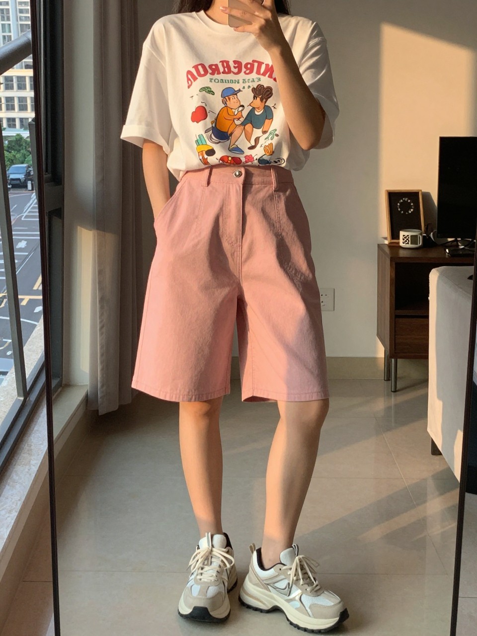 mirror selfie of a woman in pink Bermuda shorts and a graphic tee with chunky sneakers, holding phone at waist, cropped at NECK so face doesn't show, in a city apartment, evening golden hour, iPhone photo quality.