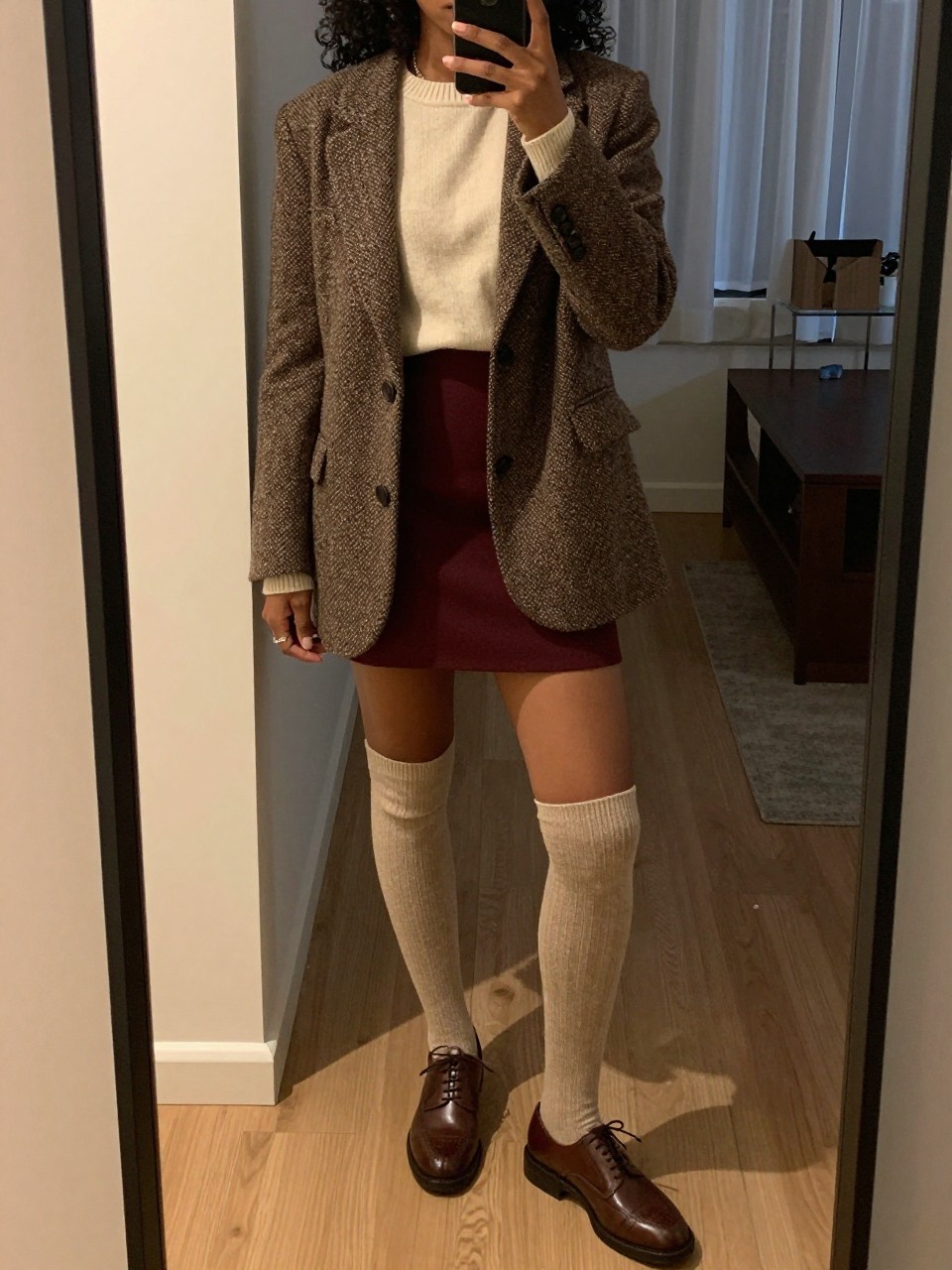 mirror selfie of a black woman wearing a brown tweed blazer over a cream sweater tucked into a maroon mini skirt with knee-high cream socks and brown oxfords, holding her phone up, cropped at NECK so face doesn't show, in a city apartment, warm moody lighting, iPhone photo quality.