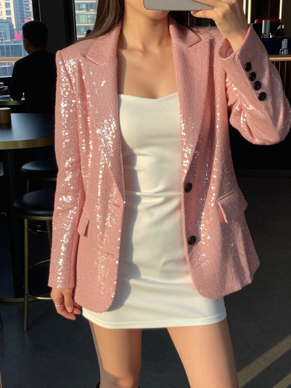 selfie of a woman in a pink sequin blazer over a white mini dress with heels, standing in a club or bar, cropped at NECK so face doesn't show, in a city apartment, evening golden hour, iPhone photo quality.