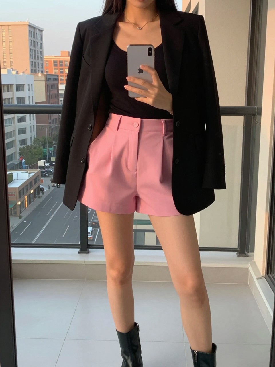 photo of a cuvy american woman in pink shorts and a black blazer over a black tank with ankle boots, holding phone at chest, cropped at NECK so face doesn't show, in a city apartment, evening golden hour, iPhone photo quality.
