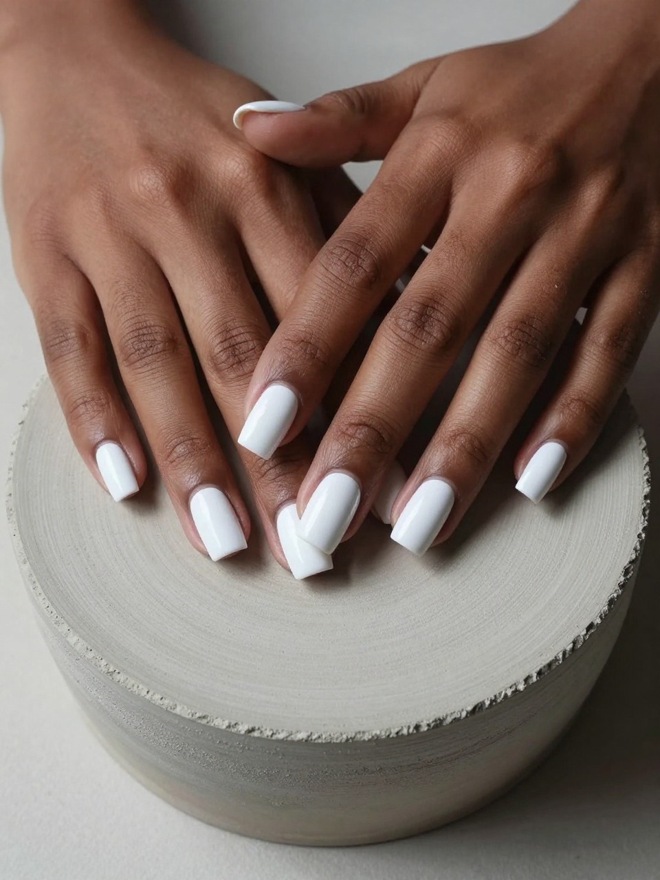 Overhead view of black feminine hands resting on a piece of raw, concrete-textured pottery. The nails are a medium square shape, coated in a pure, flat matte white. The natural half-moon shape at the base is filled with a high-gloss top coat. Angled lighting shows the dramatic difference in finish.
