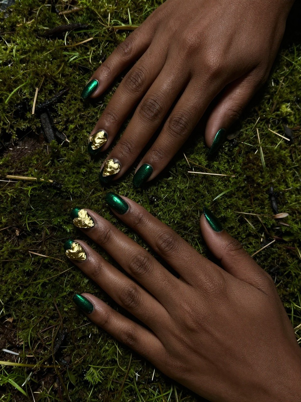 Top-down view of black feminine hands resting on a bed of moss. The nails are a deep emerald cat eye. A single, crinkled gold foil heart adorns each nail. The lighting is moody, making both the green shimmer and gold foil gleam.