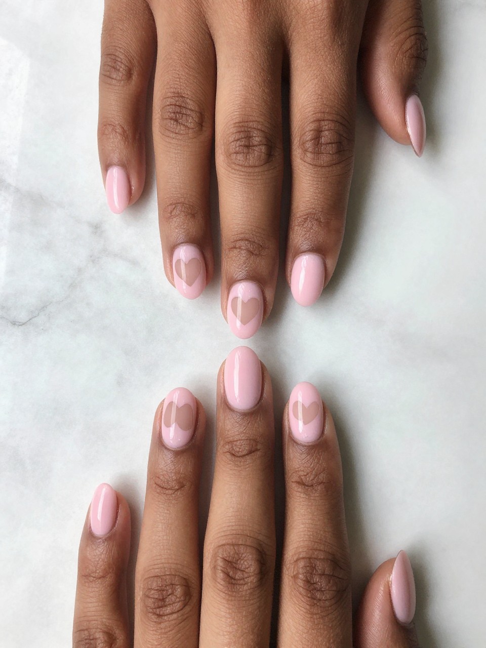 Top-down view of black feminine hands with neat, oval nails on a marble surface. The nails are a soft pink, but each has a clean, heart-shaped cut-out of bare nail at the tip. The design is architectural and intentionally cute.