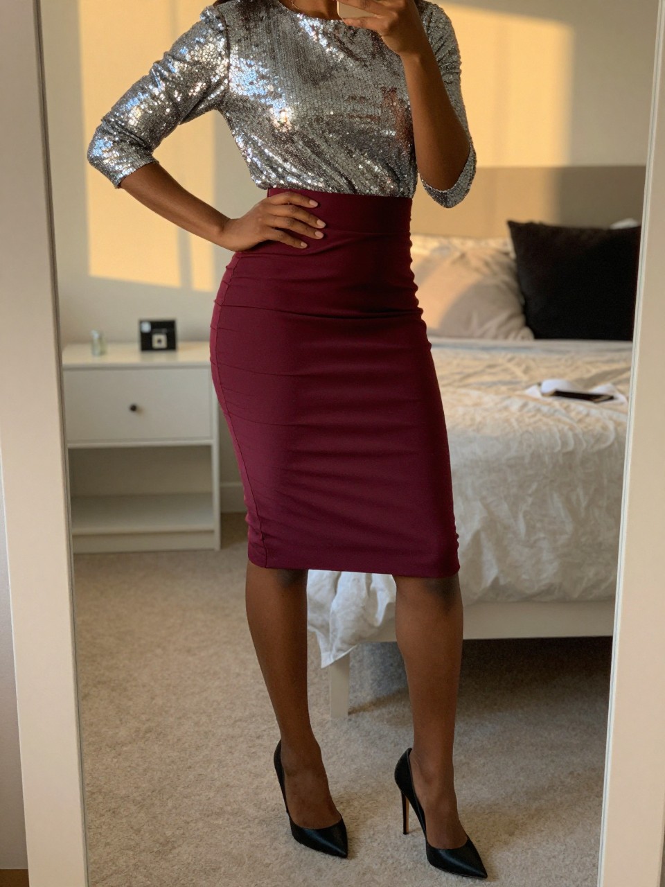 mirror selfie of a black woman wearing a silver sequin top tucked into a maroon crepe pencil skirt with black stiletto heels, one hand on her hip, cropped at NECK so face doesn't show, in a feminine-themed bedroom, evening golden hour, iPhone photo quality.