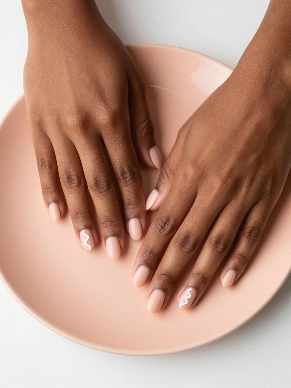 Bird's-eye view of black feminine hands with short, oval nails resting on a peach-colored ceramic plate. The nails are a matte light peach. A single, wavy white line curves across each nail. The ambiance is bright, clean, and softly feminine.