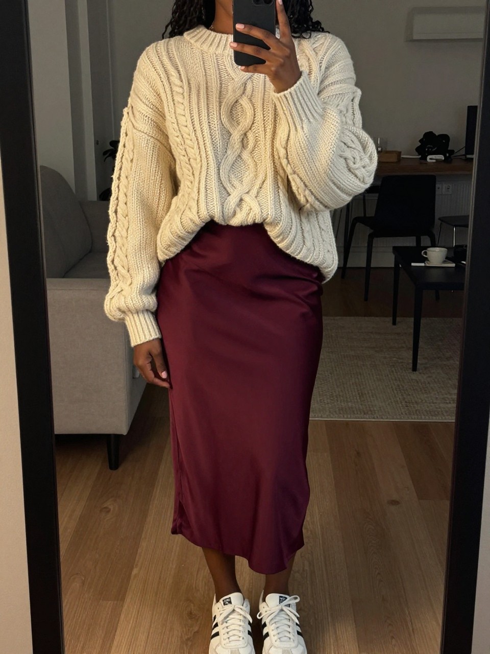 iPhone mirror selfie of a black woman wearing an oversized cream chunky cable-knit sweater half-tucked into a burgundy satin midi skirt with white sneakers, one hand holding her phone at her side, cropped at NECK so face doesn't show, in a city apartment, warm moody lighting, iPhone photo quality.